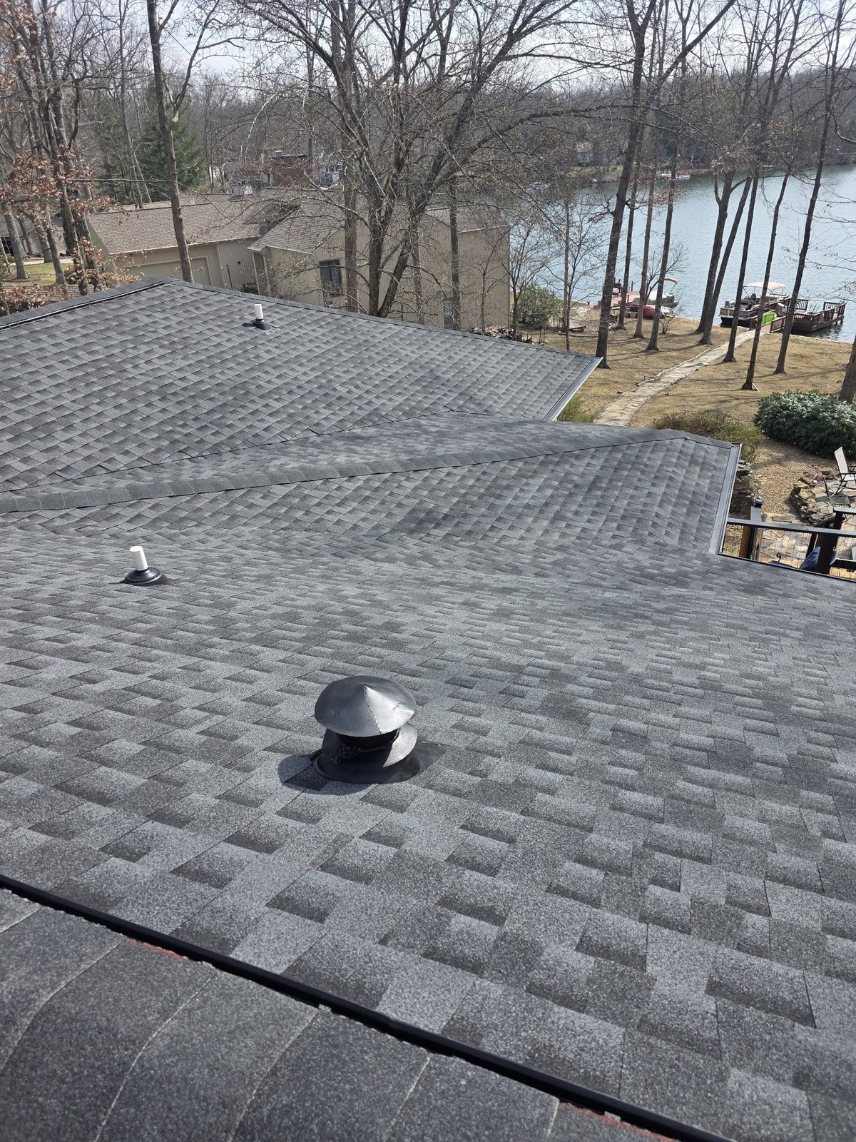 Aerial view of a gray shingled roof with ventilation caps overlooking a lake and bare winter trees