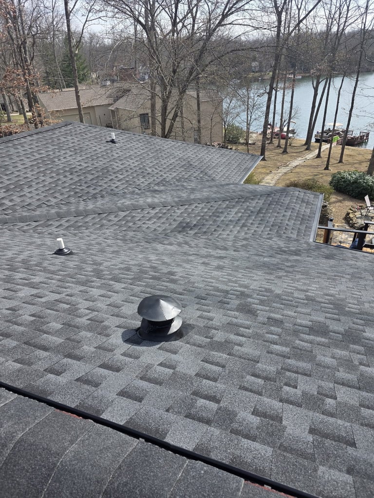 Aerial view of a gray shingled roof with ventilation caps overlooking a lake and bare winter trees