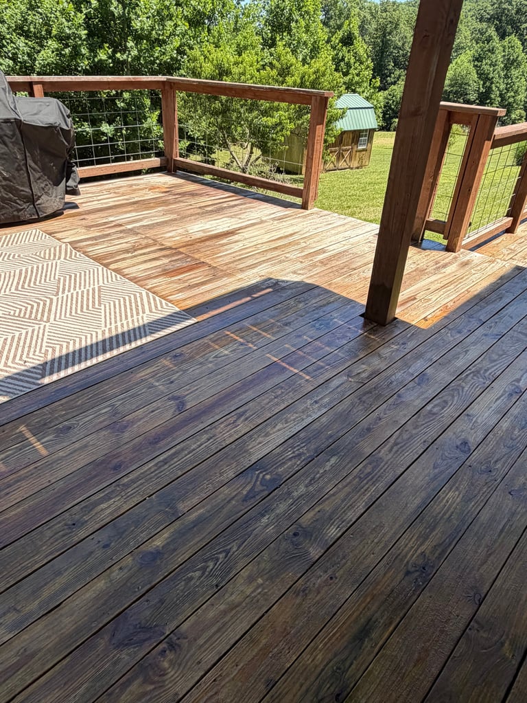 Large wooden deck with metal railings overlooking green lawn and trees, featuring stained wood flooring and supporting posts.