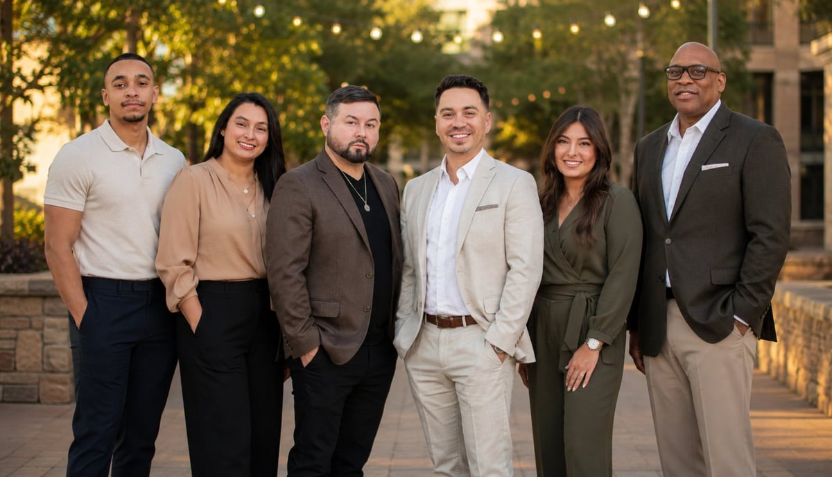 Six professionally dressed people standing together outdoors at dusk with string lights and buildings in the background