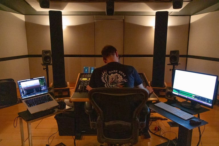 Person sitting at desk in professional recording studio with laptops, monitors, and acoustic panels on walls