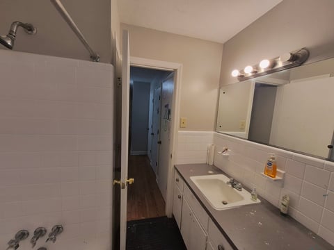 Bathroom with white subway tile, vanity sink, large mirror with vanity globe lights, and an open doorway leading back into the hallway