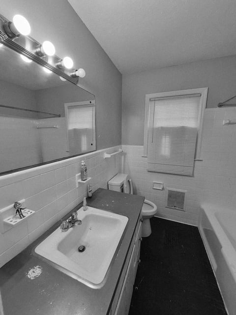 Black and white bathroom with white sink and counter, vanity mirror with globe lights, subway tile wall, window with blinds, and black tile floor