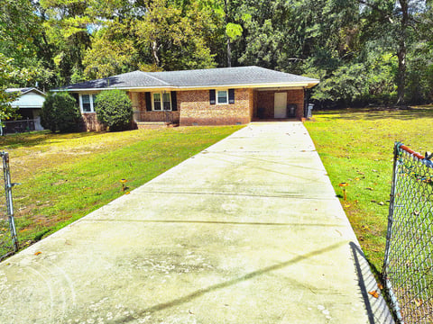 Single-story brick home with a long concrete driveway leading to the front, surrounded by green lawn and tall trees