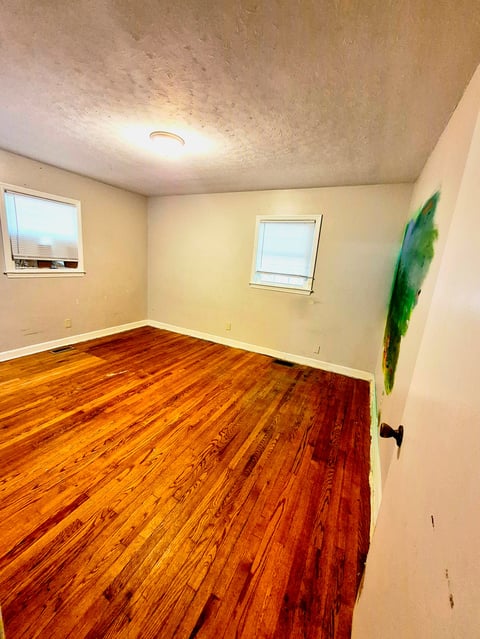 Empty room with cream walls, hardwood floor, two small windows, ceiling light, and green artwork on right wall