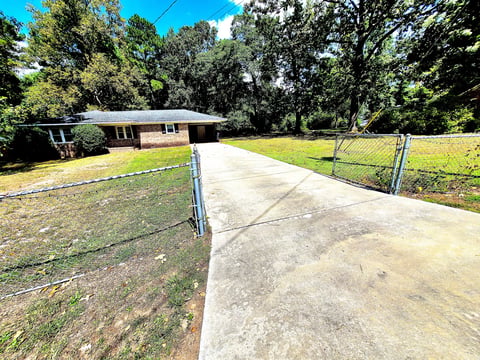 Single-story brick home with a fenced front yard, long concrete driveway, and large trees on a sunny day