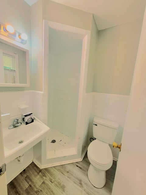 Small bathroom with white pedestal sink, toilet, glass shower enclosure, vanity mirror with lights, and wood-look flooring