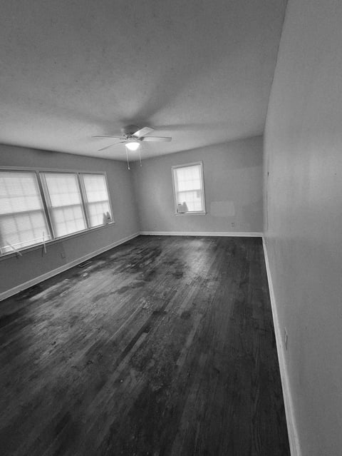 Empty bedroom with dark hardwood floors, white walls, ceiling fan, and windows with natural light in black and white