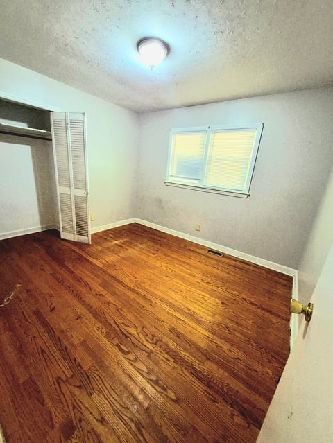 Empty bedroom with hardwood floors, louvered closet doors, a window with blinds, and a ceiling light fixture