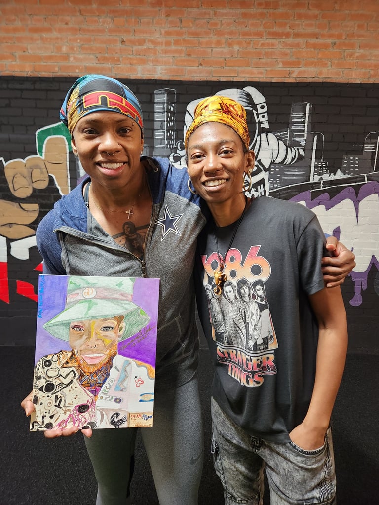 Two smiling people posing together in front of a brick wall and street art mural, one holding a colorful portrait artwork