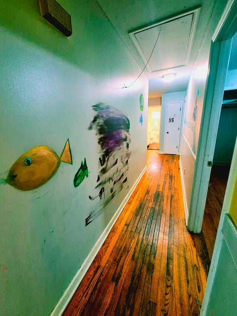 Hallway with hand-painted fish murals in yellow, green, and blue on the left wall, hardwood floors, and the white door numbered 55 visible at the end