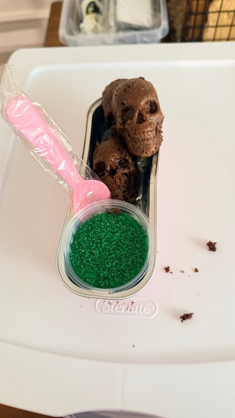 Chocolate skull-shaped candy with pink wrapper next to a container of green sugar granules on a white surface