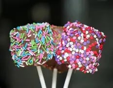 Three colorful decorated cake pops on sticks - one with rainbow sprinkles, one with pink and white pearls, and one with red sprinkles