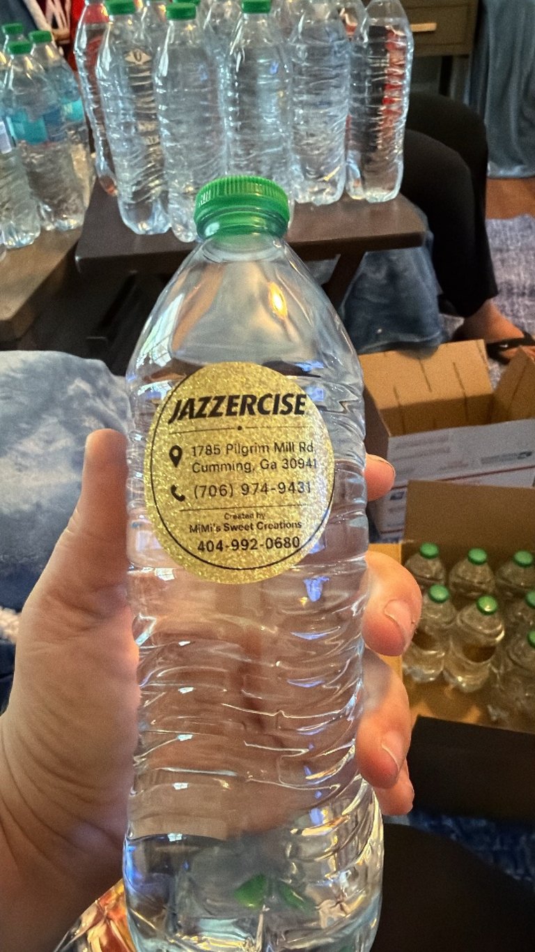 Hand holding a clear plastic water bottle with a green cap and gold Jazzercise label, surrounded by other bottles