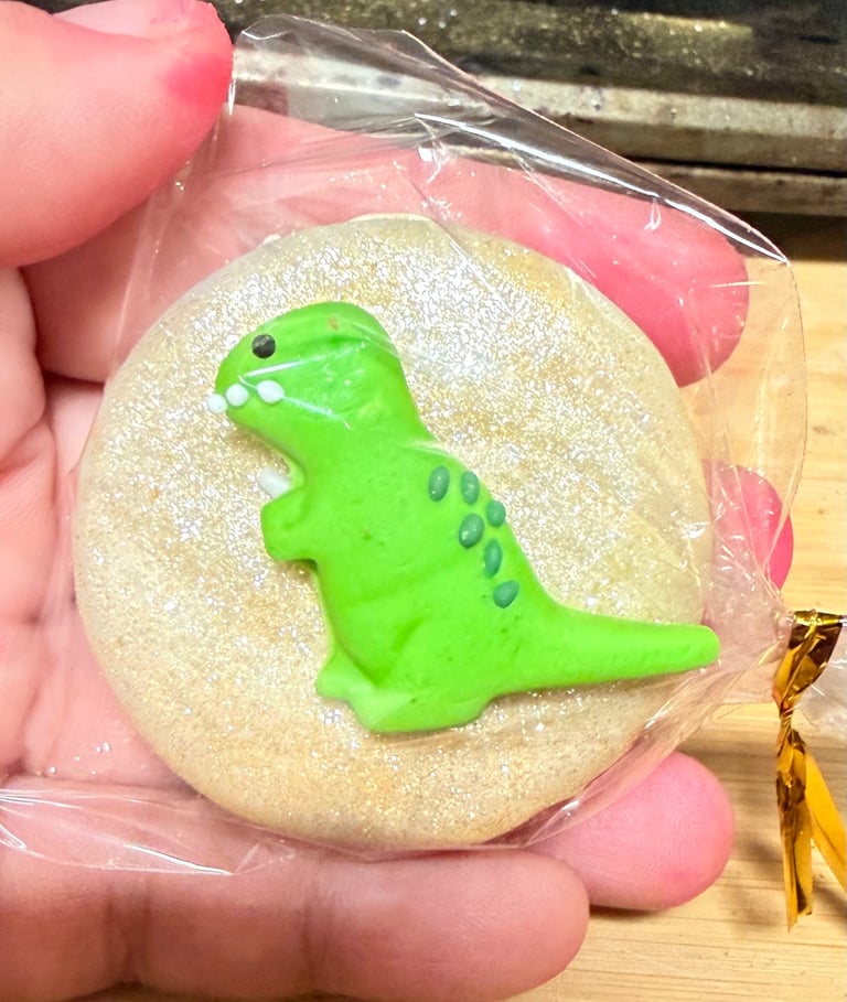 Hand holding a round sugar cookie decorated with gold glitter and a bright green dinosaur topper, wrapped in clear cellophane with pink tissue paper