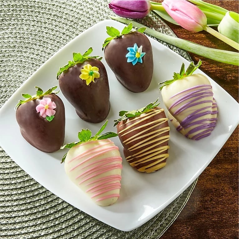 Decorated chocolate-covered strawberries on a white plate with colorful icing drizzles and fondant flowers, displayed on a green cloth napkin with tulips