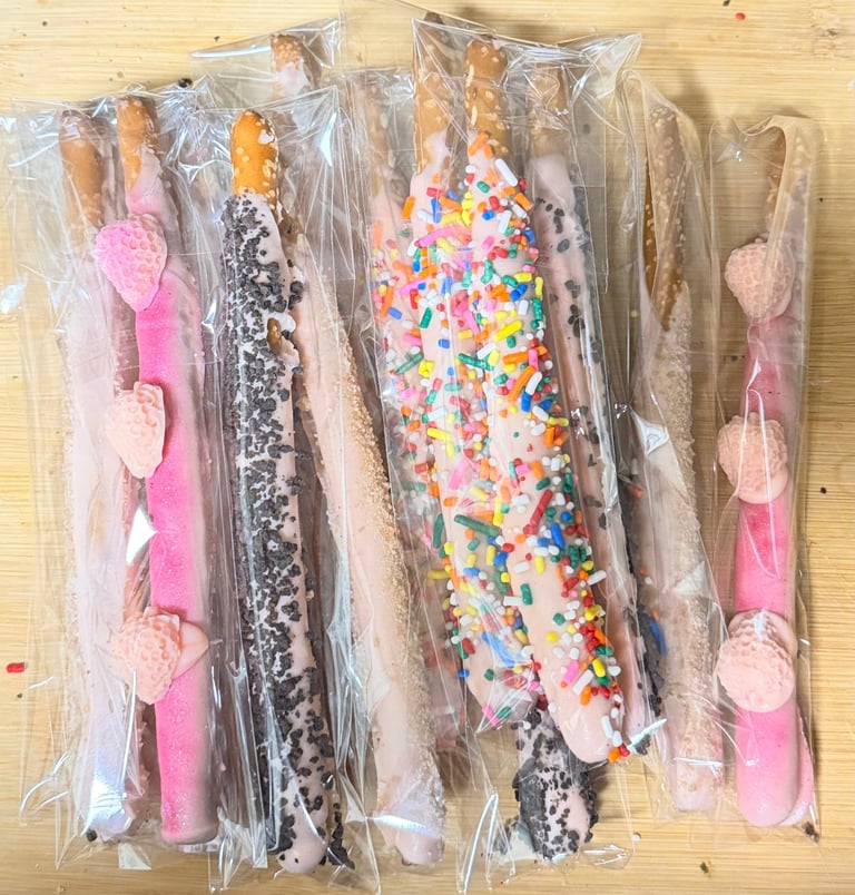 Five individually wrapped chocolate-covered pretzel rods with various toppings including sprinkles, coconut, and colorful candies on a wooden surface