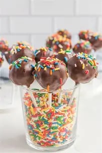 Chocolate cake pops with colorful sprinkles displayed in a glass filled with rainbow sprinkles