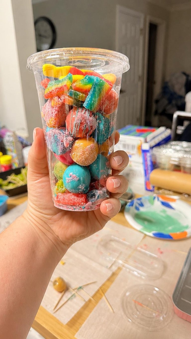 Hand holding a clear plastic cup filled with colorful sour gummy candies and round gumballs in various colors including red, yellow, blue, and green.
