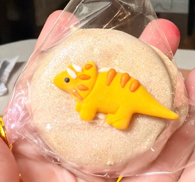 Yellow dinosaur toy on a round beige cookie or cake with pink wrapping, held in someone's hand