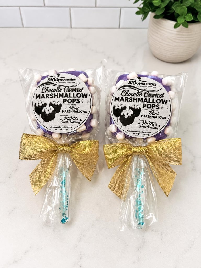 Two chocolate-covered marshmallow pops with gold ribbons and blue sticks on a marble counter with a potted plant.