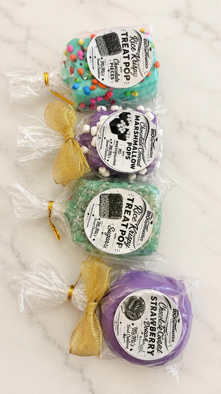 Four colorful bath bombs in clear plastic packaging with white labels and gold ribbon bows, arranged vertically on a marble surface.