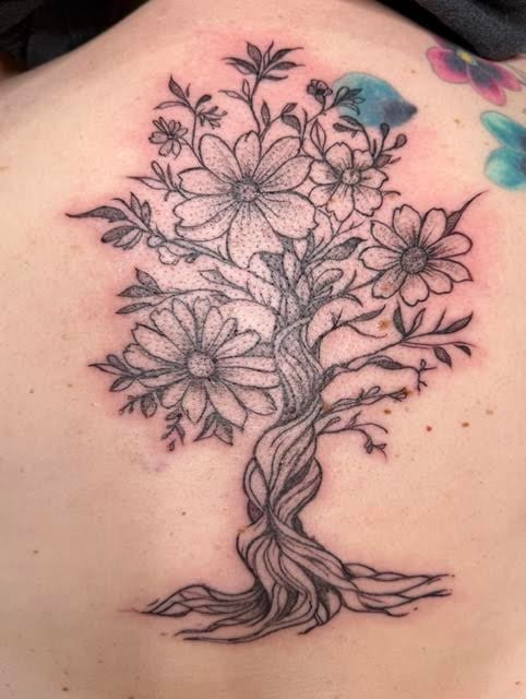 Black ink tattoo of a flowering tree with daisy-like blooms and twisted trunk on pale skin, with colorful watercolor accents visible above