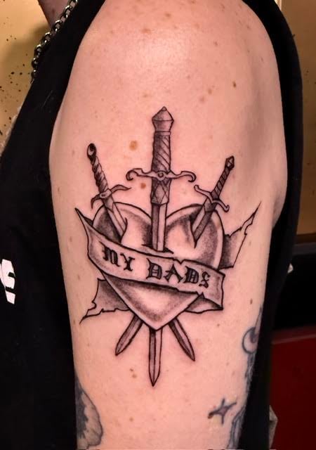 Black ink tattoo on arm featuring a heart shield with swords and text reading my dame