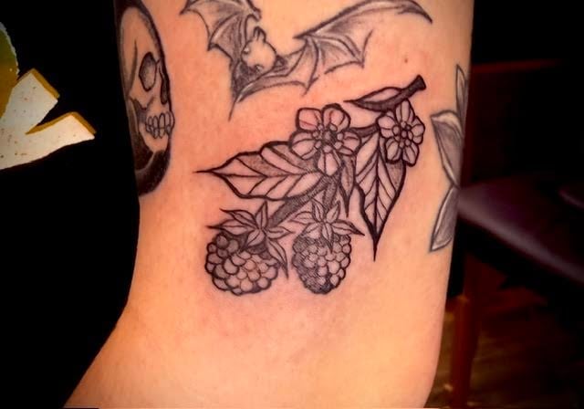 Arm tattoo featuring blackberry branches with detailed berries and leaves, along with bat and skull designs