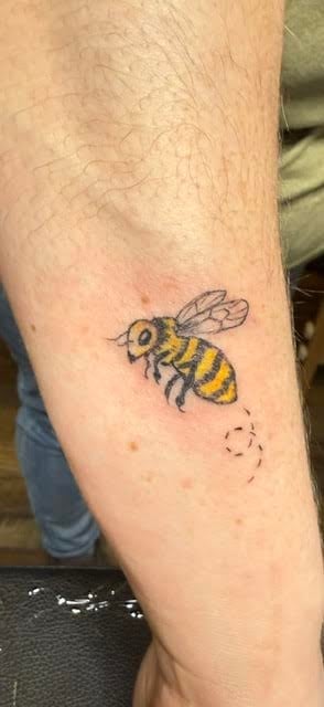 A small bee tattoo on someones arm with yellow and black stripes and a dotted trail beneath it