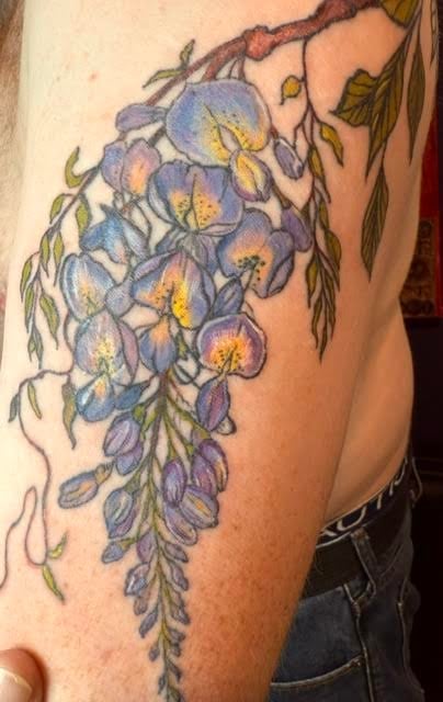 Tattoo of cascading blue and purple wisteria flowers with green leaves on upper arm