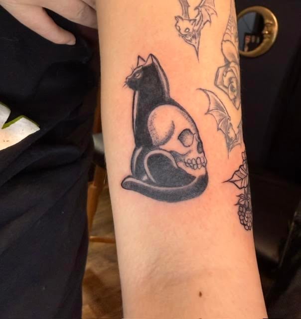 Black cat tattoo holding a skull, positioned on persons forearm next to other tattoos