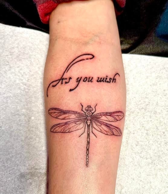 Forearm tattoo featuring cursive text reading As you wish with a dragonfly illustration below it in black ink