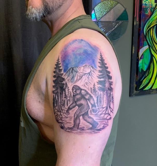 Man displaying a shoulder tattoo of a Bigfoot figure in a forest landscape with mountain and blue moon design