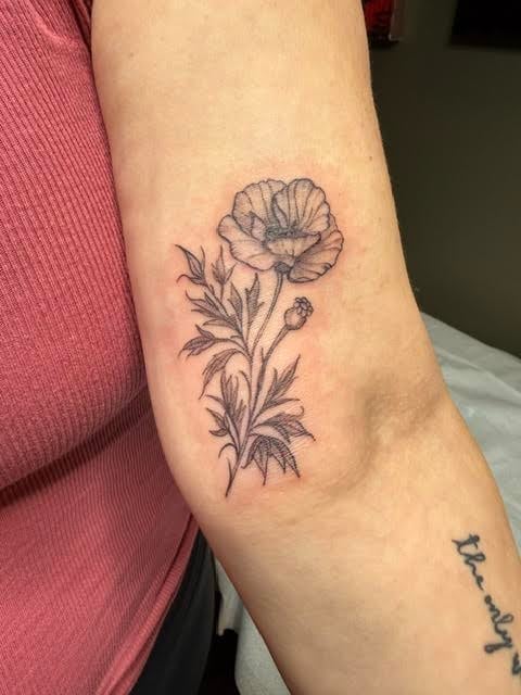 Black outline tattoo of a poppy flower with stem and leaves on an arm, worn by a person in a pink shirt