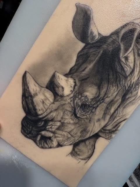 Black and white detailed tattoo of a rhino head on skin, showing realistic shading and facial features