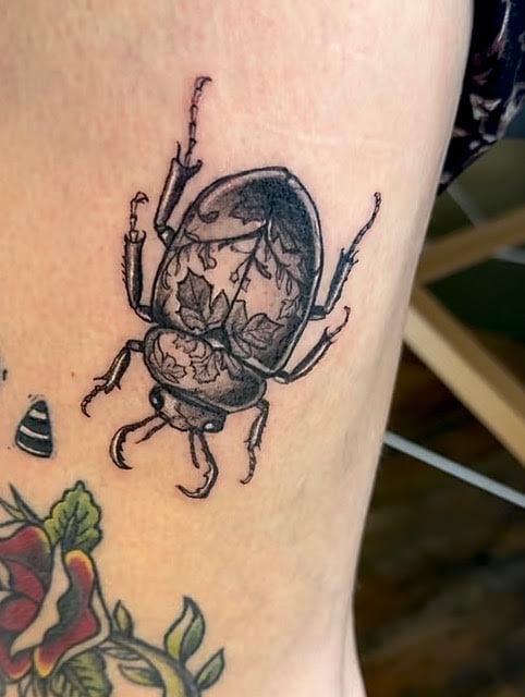 Tattoo of a detailed black ink beetle with ornate wing patterns and antennae on skin with colored floral design below