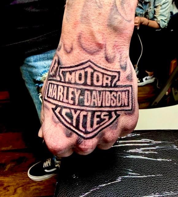 Hand tattoo displaying Harley-Davidson Motor Cycles logo in black ink on the back of someones hand