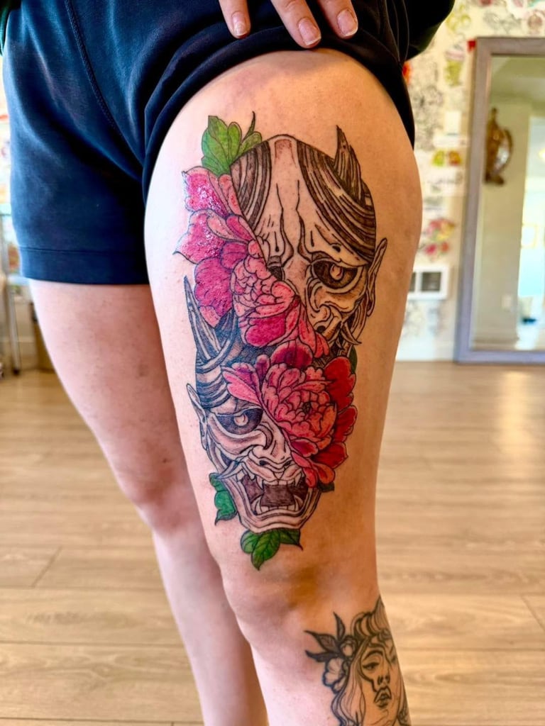Tattoo on persons thigh featuring ornate demon mask with pink and red flowers intertwined in artistic design