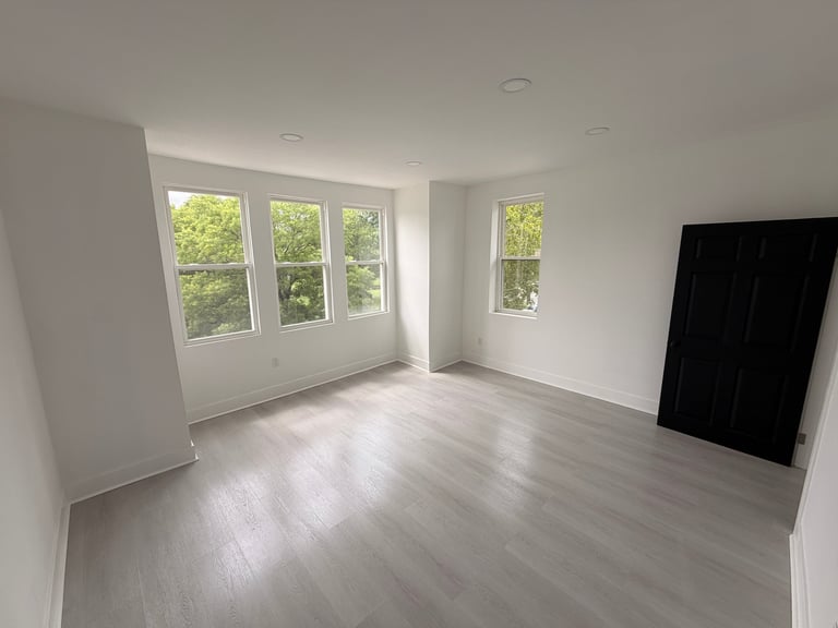 Bright empty room with light wood floors, white walls, large windows with trees visible outside, and recessed ceiling lights