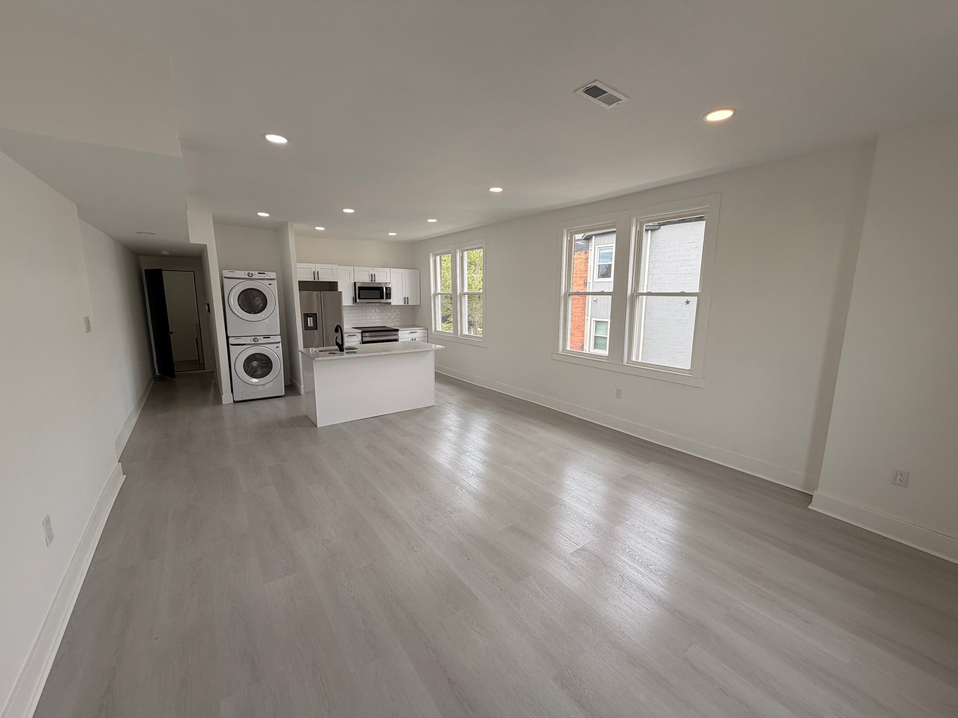 Modern open-concept apartment interior with gray wood floors, white walls, stacked washer-dryer, kitchen island, and large windows