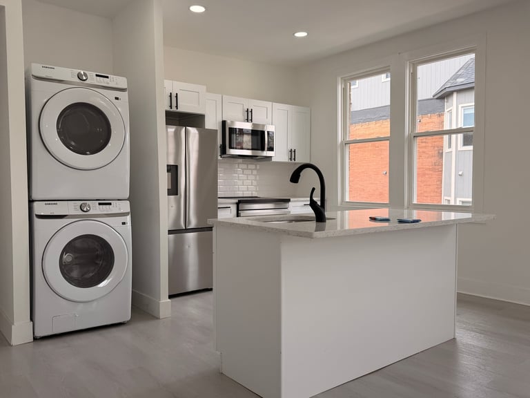 Modern laundry room with stacked white washers and dryers on left, white cabinets, stainless steel appliances, island counter, and large windows with brick exterior view