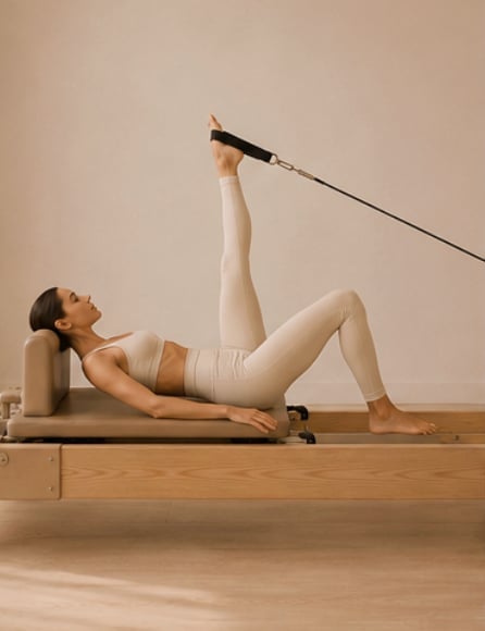 Woman performing pilates leg extension exercise on reformer machine with beige walls and wooden flooring
