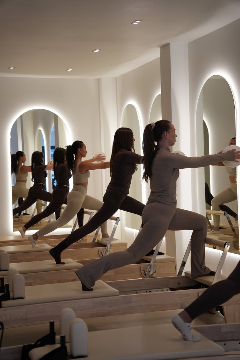 Multiple people performing a Pilates exercise on reformer machines in a modern, brightly lit studio with mirrored walls