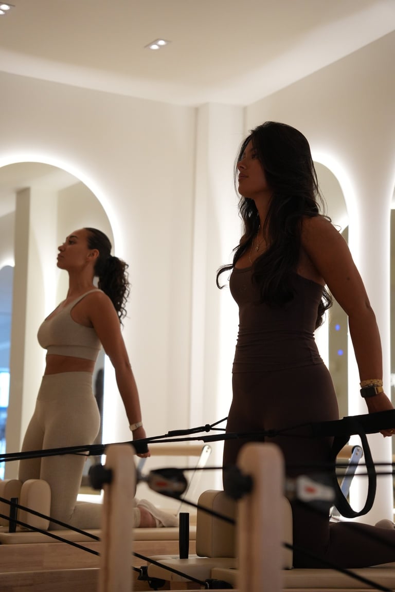 Two women in a modern dressing room with illuminated mirrors, one adjusting athletic wear while a bicycle is visible in the foreground.