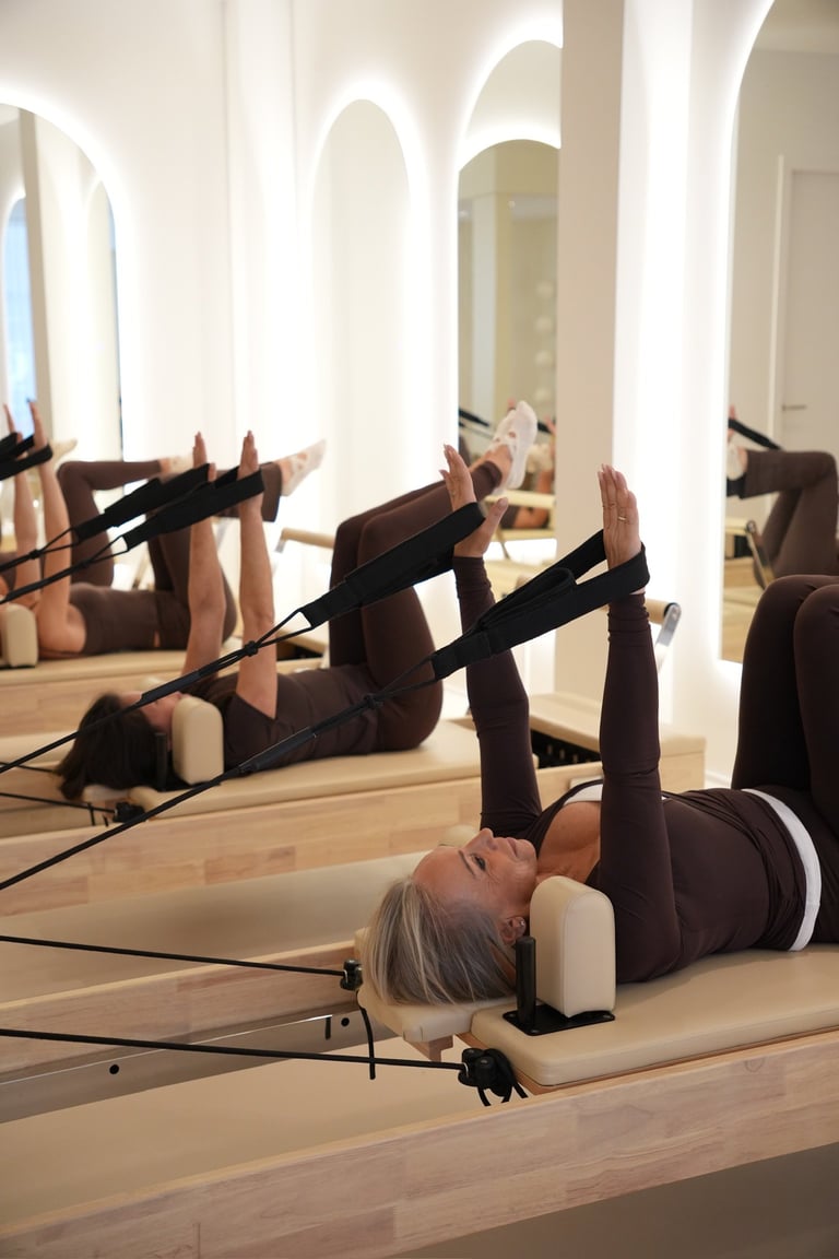 Group fitness class performing pilates exercises on reformer machines in a bright, mirrored studio with white columns