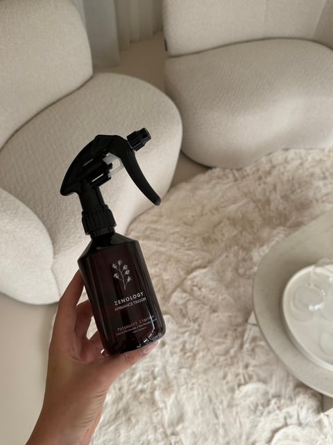 Hand houdt een donkerbruine Mixology ambiance spray fles vast tegen beige fauteuil en kussens