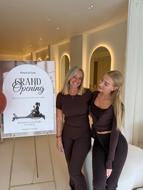 Twee vrouwen in zwarte outfits poseren naast het House of Core Grand Opening bord