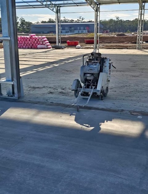 Concrete cutting machine on warehouse floor with pink insulation rolls and construction materials visible in background