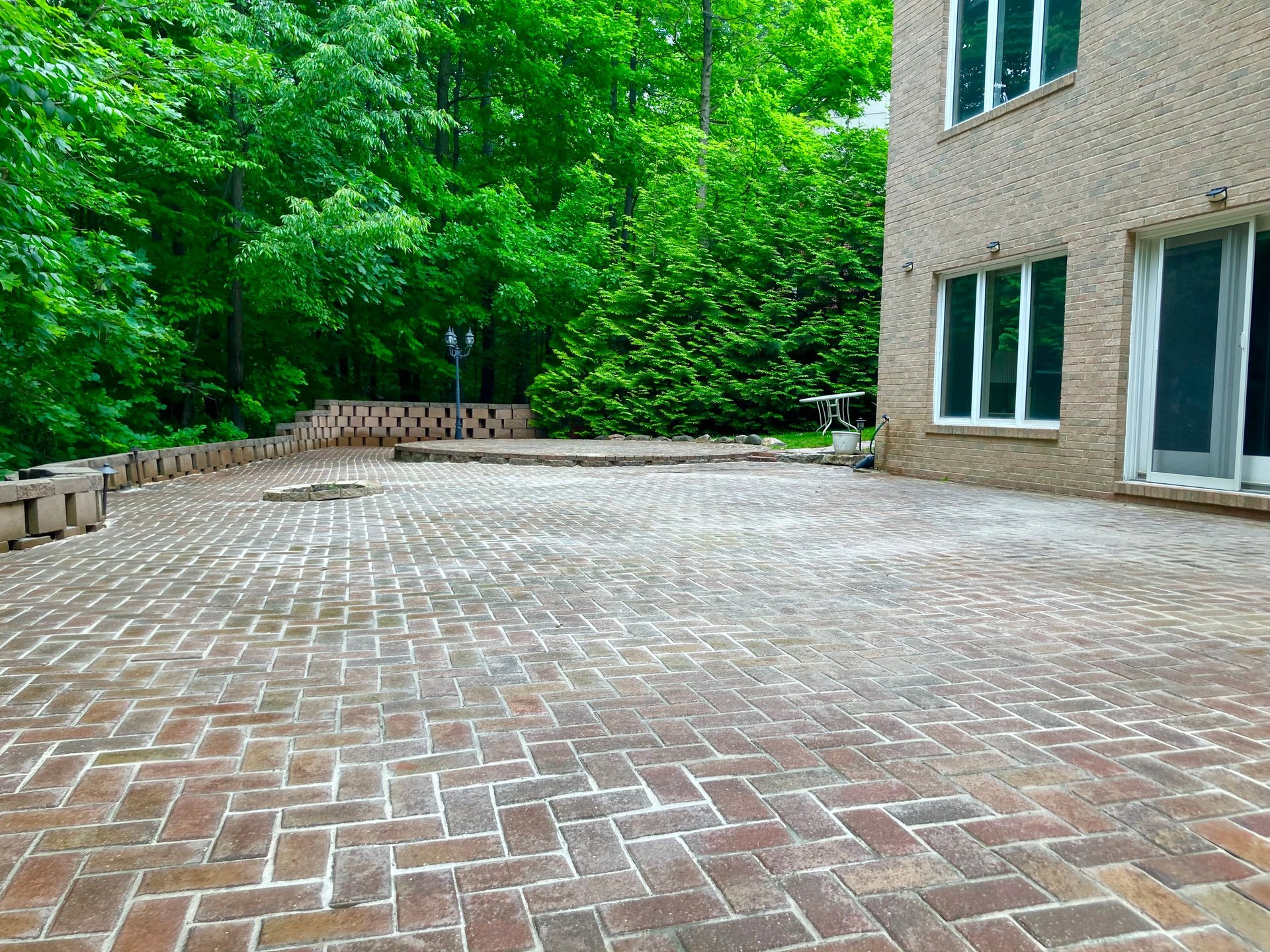 Expansive brick patio with herringbone pattern beside modern brick house surrounded by dense green foliage